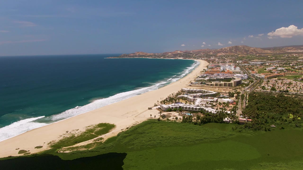 Wide aerial frame of San Jose Del Cabo coastline with white waves running along the sand, a green foreground area, clustered coastal buildings, distant hills, and no visible people.