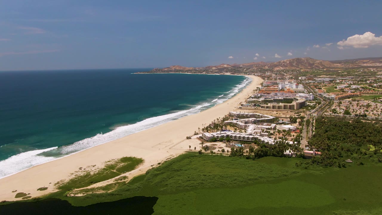 Aerial view of San Jose Del Cabo beach with the ocean occupying the left side, white surf tracing the shore, developed buildings near the sand, green land in front, and no visible people.