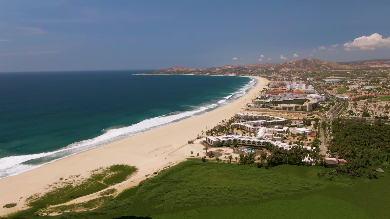 Wide aerial view over San Jose Del Cabo, Mexico, with blue ocean on the left, a long sandy beach, green foreground vegetation, coastal buildings, inland hills, and no clearly visible people.