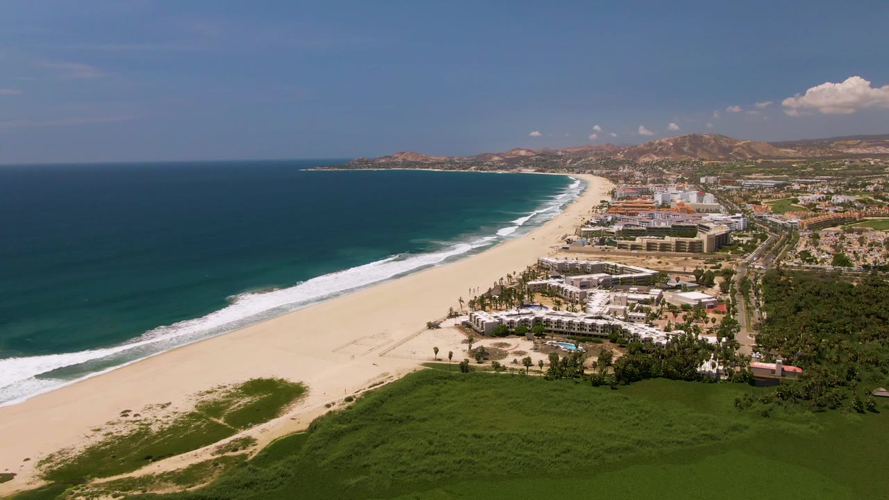 Drone view along a sandy shoreline with waves, beach buildings, and hills beyond.