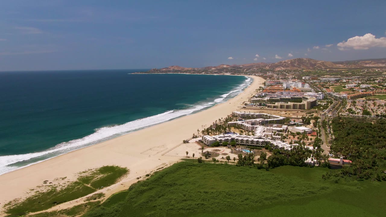 San Jose Del Cabo aerial view showing dark blue water, a bright strip of beach, white surf lines, green vegetation in the lower frame, coastal buildings to the right, and low hills in the distance.