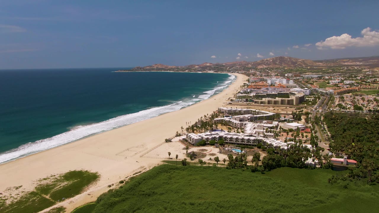 Aerial frame over San Jose Del Cabo, Mexico showing a wide sandy beach beside blue ocean, resort buildings, green vegetation, hills, and tiny beach walkers near the waterline.