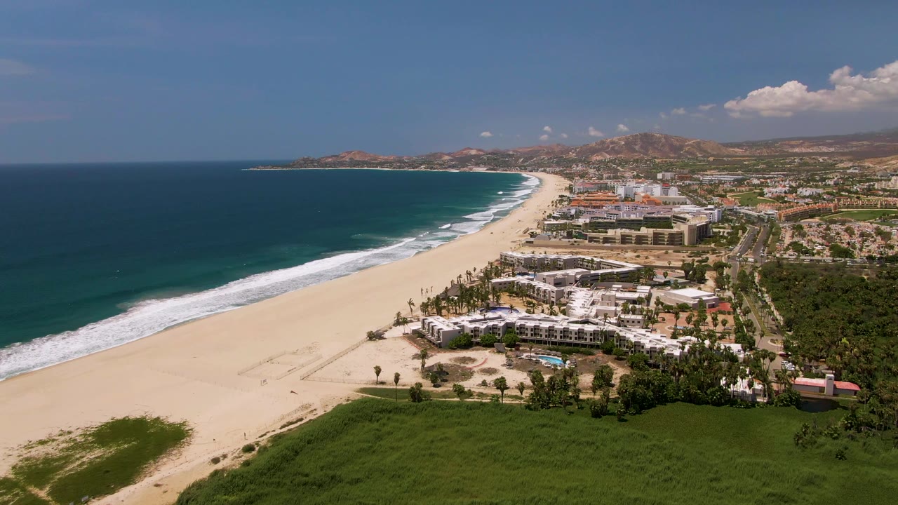 Aerial view along the San Jose Del Cabo beach with white surf bands, open sand, a resort district to the right, green vegetation at the bottom of frame, and dry hills beyond town.