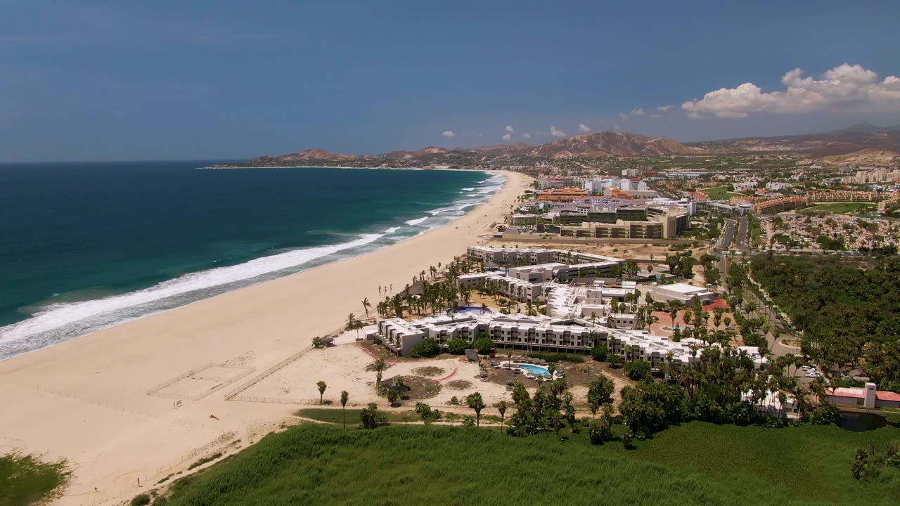 A high aerial view over San Jose Del Cabo shows a wide sandy beach, blue ocean surf, resort buildings, palms, roads, and distant hills, with no visible people.