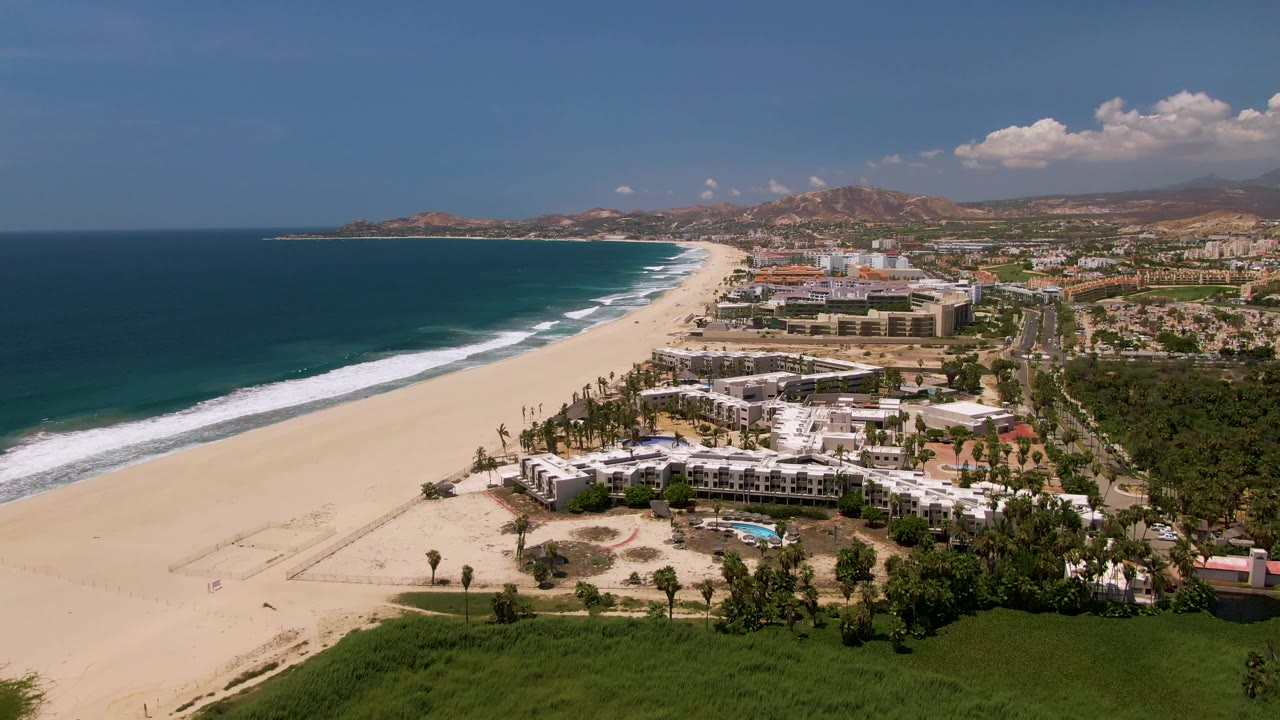 The aerial frame looks along the San Jose Del Cabo shoreline, with the beach and surf stretching left of a resort complex and town buildings under scattered clouds, with no visible people.