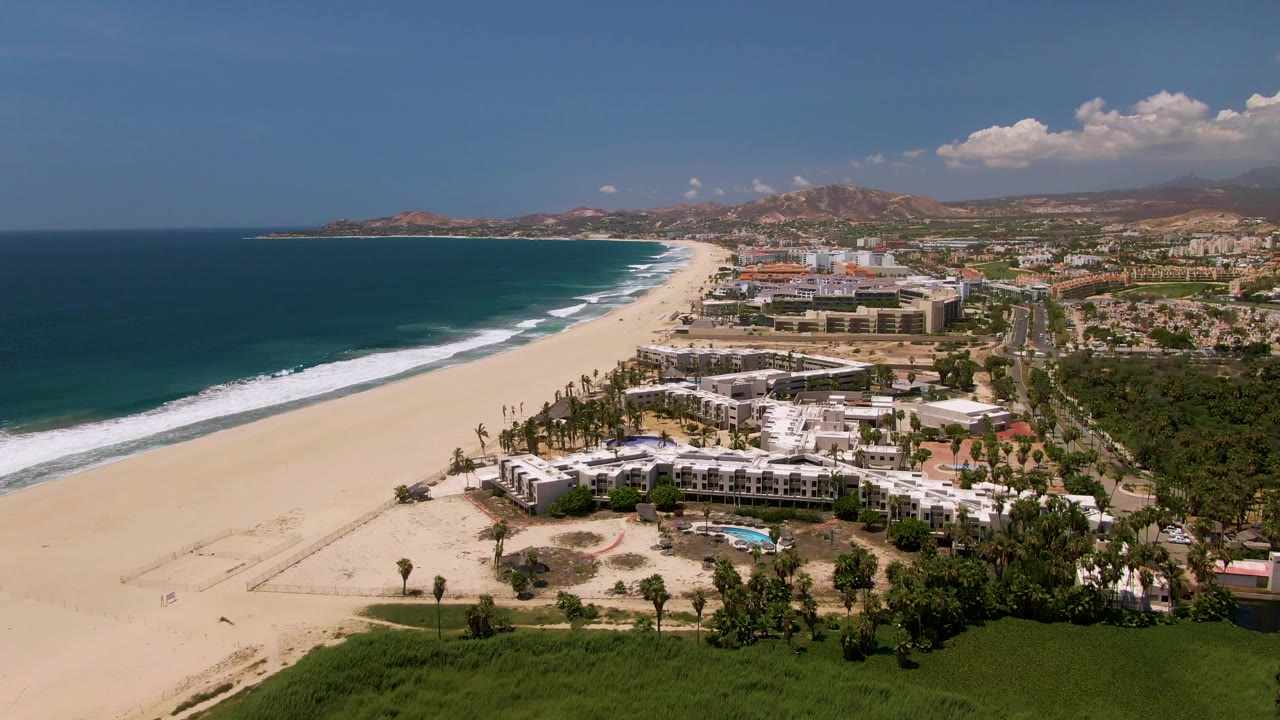 A wide San Jose Del Cabo coastal aerial shows pale sand, dark blue water, white wave bands, low resort buildings, a pool area, palms, and hazy hills in the background, with no visible people.