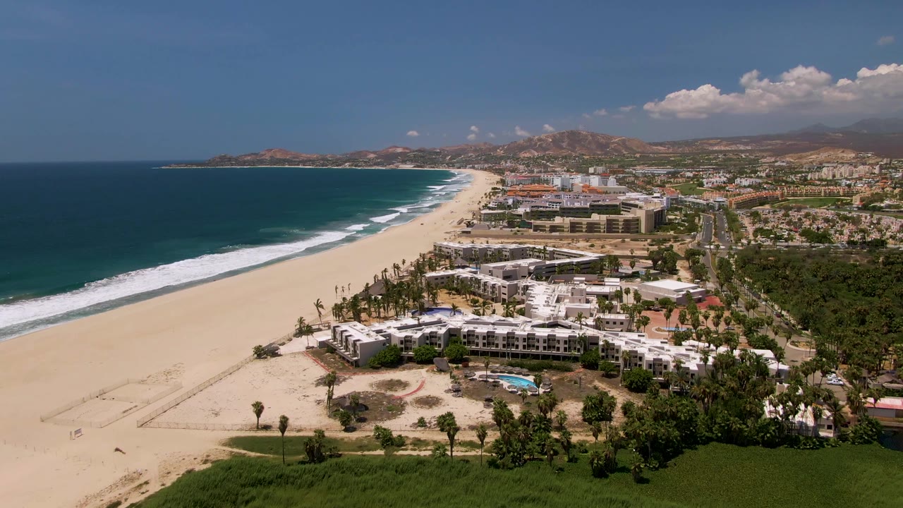 Aerial coastline view with open beach, resort roofs, palms, and ocean.