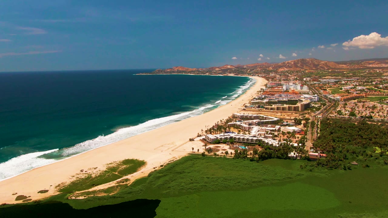 Aerial view of sandy beach, ocean waves, coastal buildings, green foreground, and distant mountains.