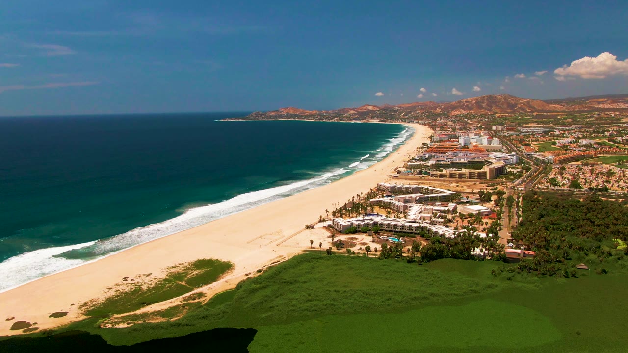 Second frame of the San Jose Del Cabo coastline shows the surf line slightly farther along the beach, with ocean on the left, buildings at center right, green foreground, and no visible people.