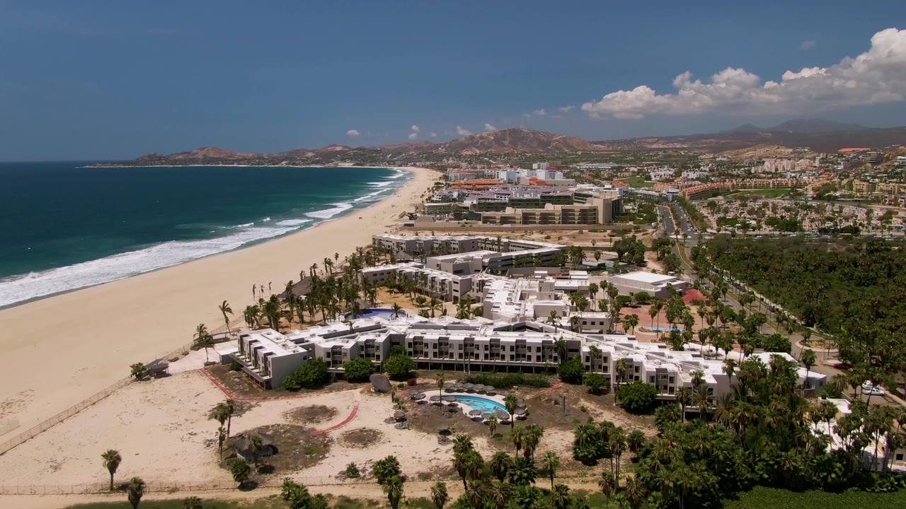 Wide aerial frame of San Jose del Cabo beachfront resorts with surf bands along the left shoreline, white hotel blocks near the sand, a small pool below, and no visible people.
