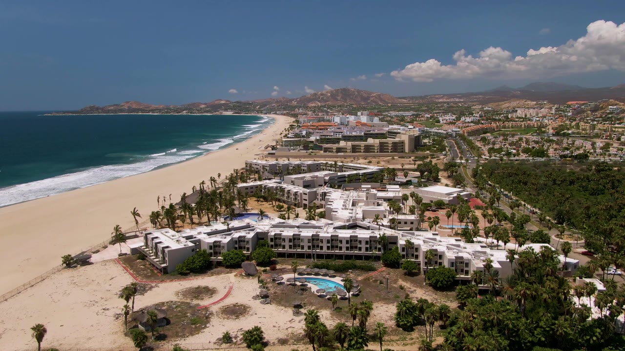 Aerial view in San Jose del Cabo showing a beachfront resort complex, ocean waves breaking along the sand, a pool in the lower foreground, hazy hills behind town, and no visible people.
