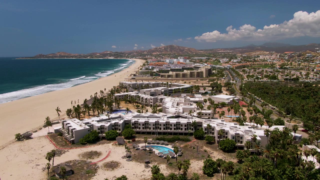 San Jose del Cabo aerial frame with the beach and ocean running diagonally left, resort buildings across the center, a red barrier visible on the sand in the foreground, and no visible people.