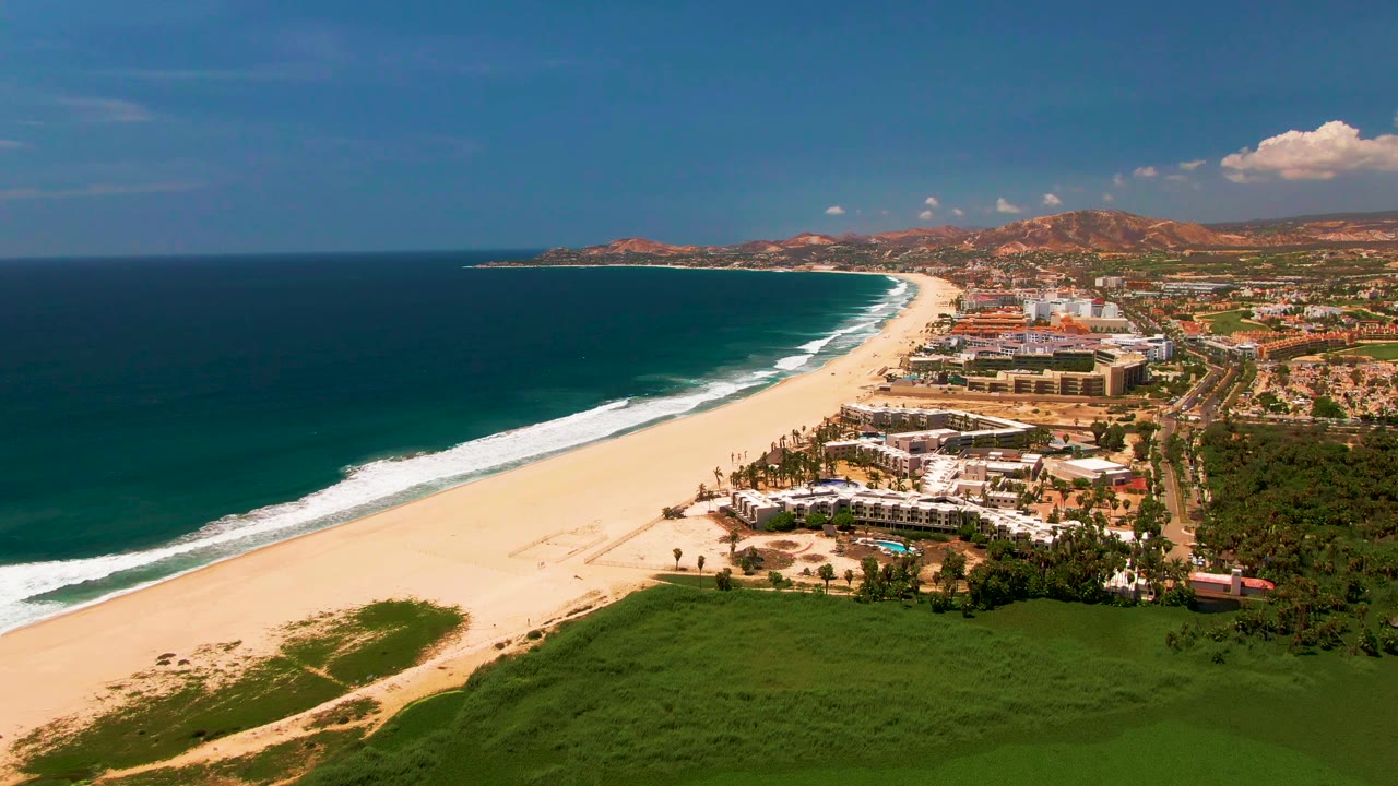 Opening aerial frame over San Jose del Cabo shows dark blue ocean, a long sandy beach, white surf, resort buildings, green foreground vegetation, distant hills, and no visible people.