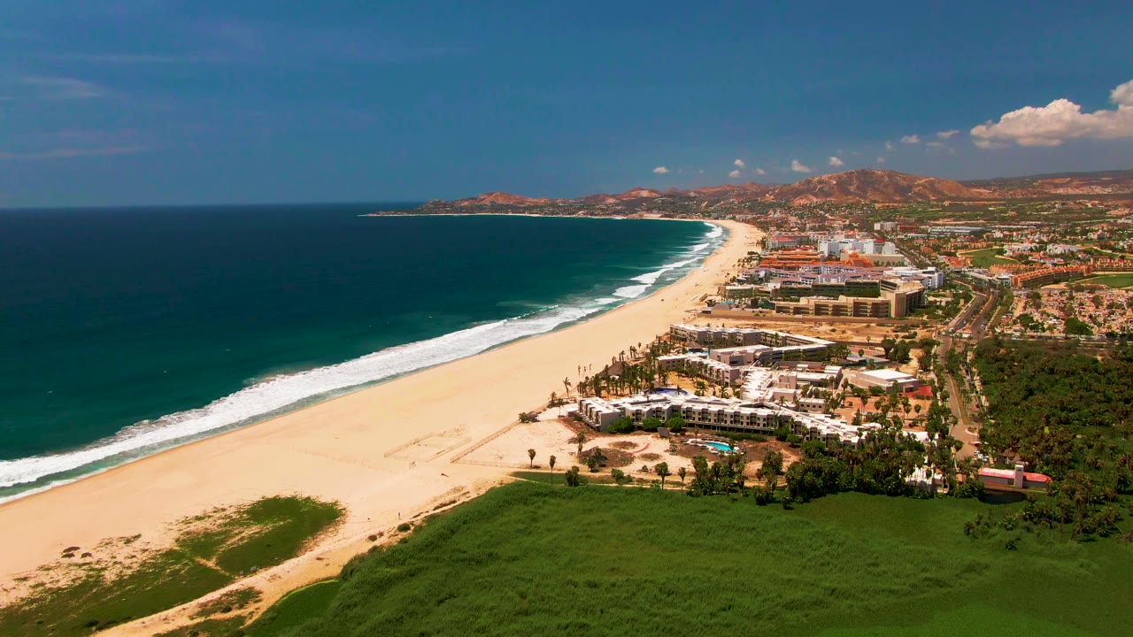 At 0:02, the aerial view shows the curved San Jose del Cabo beach between blue ocean and inland resort buildings, with a green foreground strip, brown hills in the distance, and no visible people.