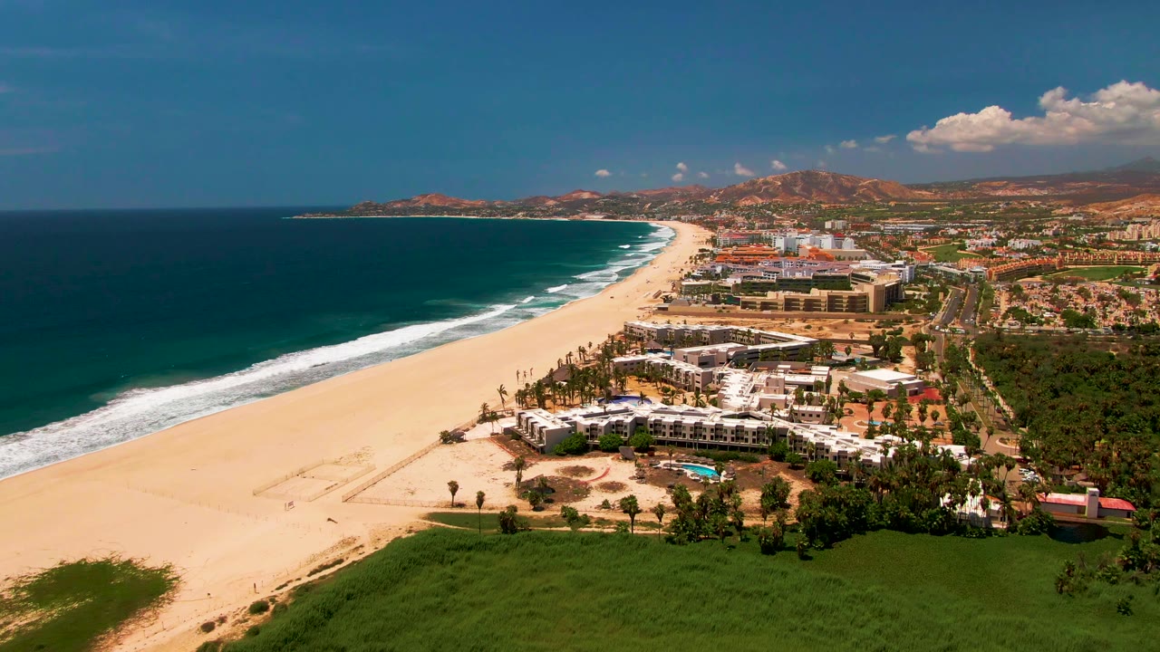 Aerial view over San Jose del Cabo with a wide sandy beach, dark blue ocean, white surf lines, coastal buildings, green foreground vegetation, inland hills, and tiny visible people on the sand.