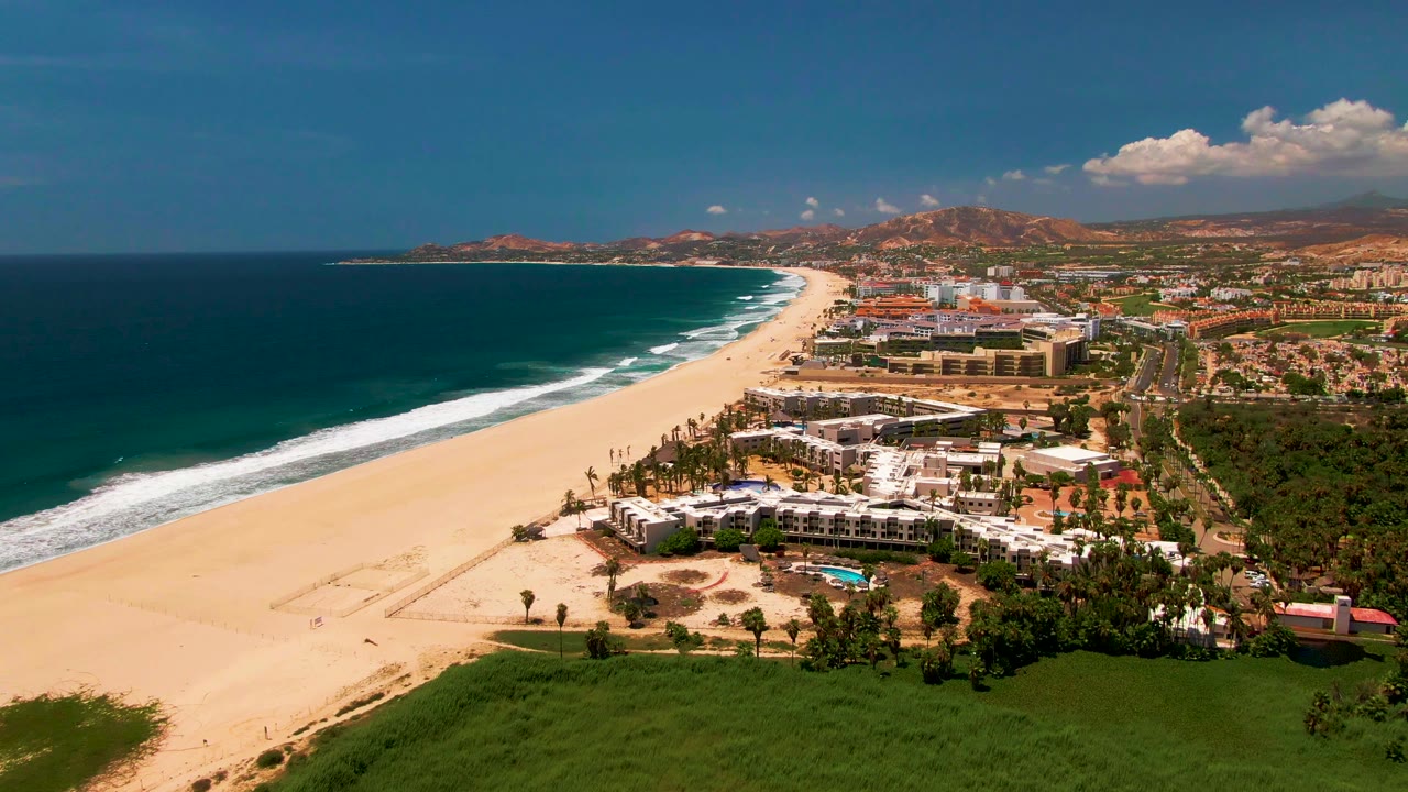 Wide drone view of beach surf beside resort-area buildings and inland hills.