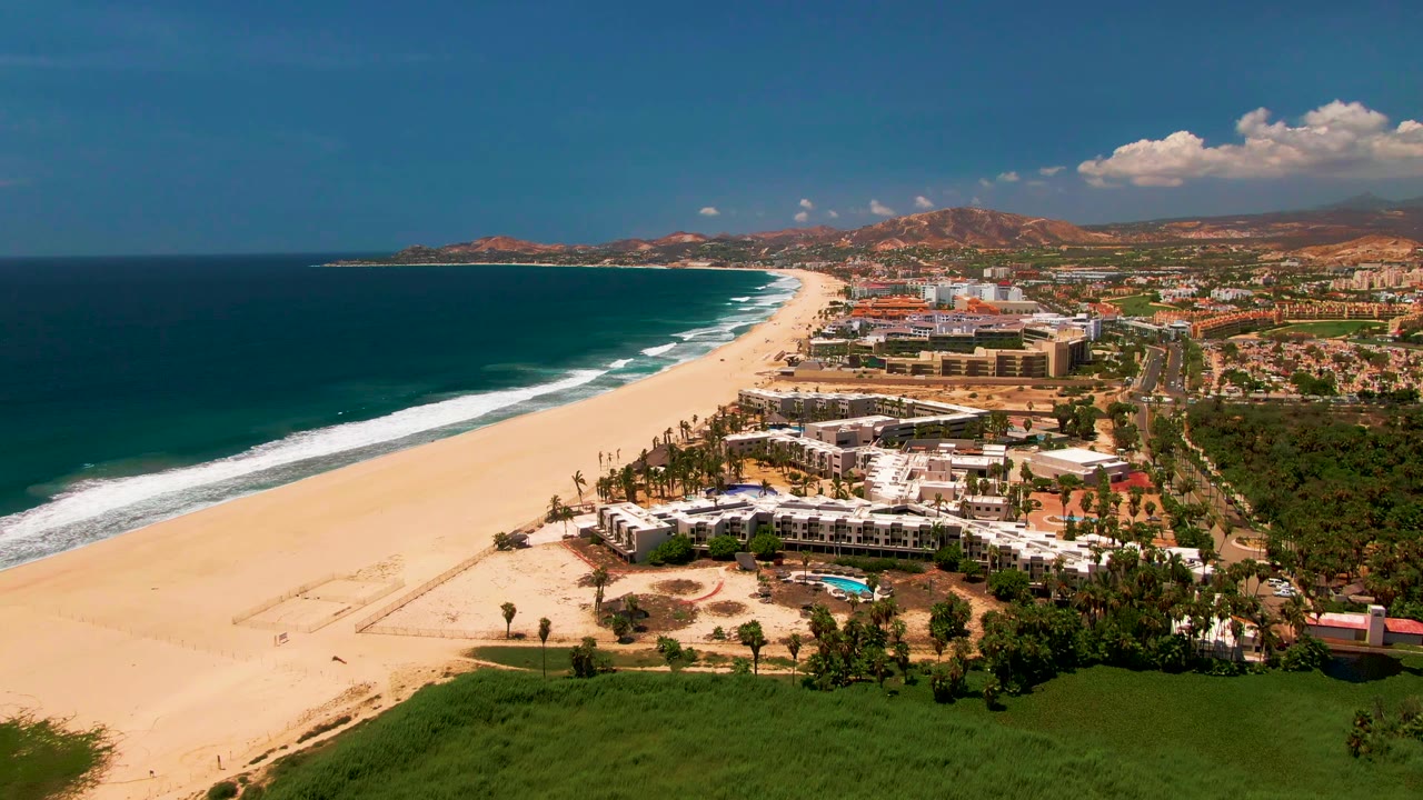 San Jose del Cabo aerial coastline with a broad pale beach, darker ocean water, white surf breaking close to shore, clustered coastal buildings, palm trees, green vegetation, hills, and visible small beachgoers.