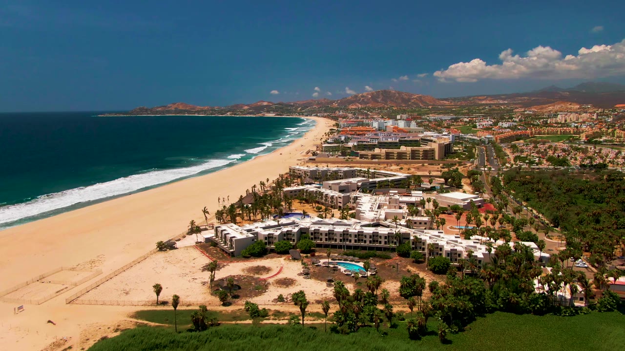 Opening aerial frame over San Jose Del Cabo showing a long sandy beach, blue ocean, white surf, a beachfront building complex, inland roads, low mountains, and no visible people.