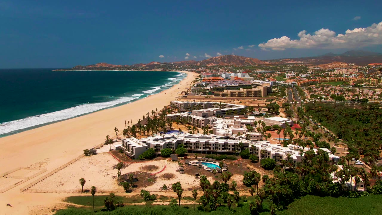 Wide drone view of beach, ocean surf, resort buildings, and a white structure near the shore.