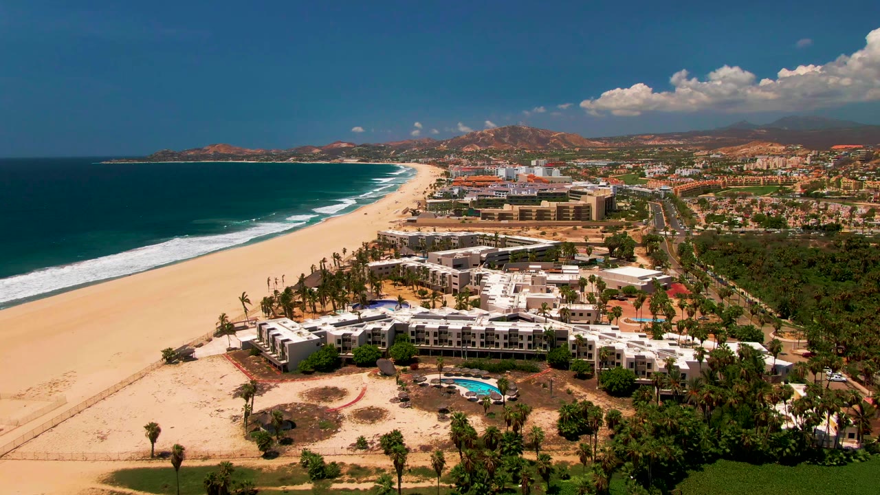 Third San Jose Del Cabo aerial frame showing the beach and ocean stretching into the distance, the white construction area beside palms, inland roads, hills, clouds, and no visible people.