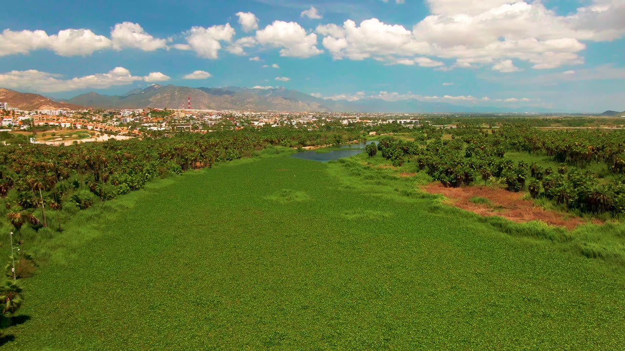 Aerial frame in San Jose del Cabo showing the green field stretching toward tree cover, a small water channel, distant buildings, mountain ridges, bright clouds, and no visible people.