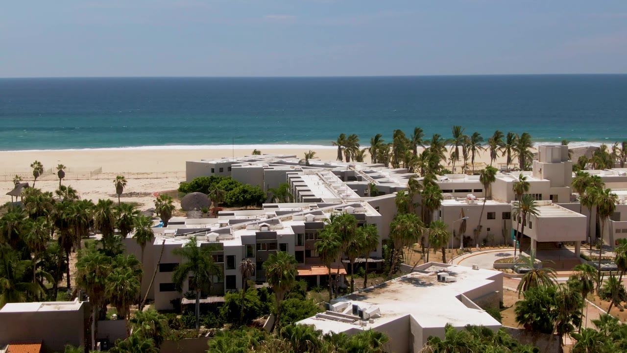 Aerial frame in San Jose del Cabo showing white resort rooftops and palms in front of a sandy beach and blue ocean, with no visible people.