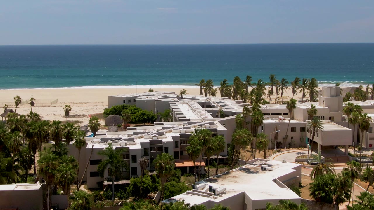 High drone view in San Jose del Cabo with resort rooftops filling the lower frame, palm trees along the beach, and a clear blue ocean horizon, with no visible people.
