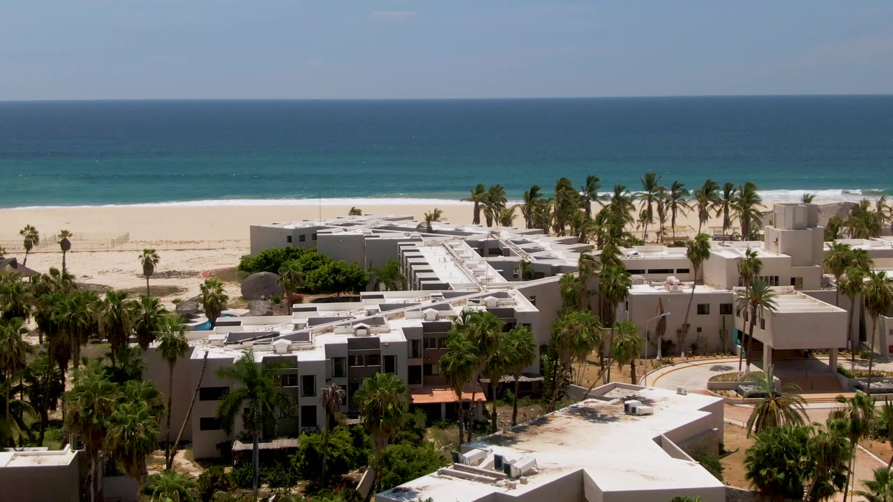 San Jose del Cabo aerial frame with low-rise beachfront buildings, clustered palms, pale sand, and blue-green ocean bands in the distance, with no visible people.