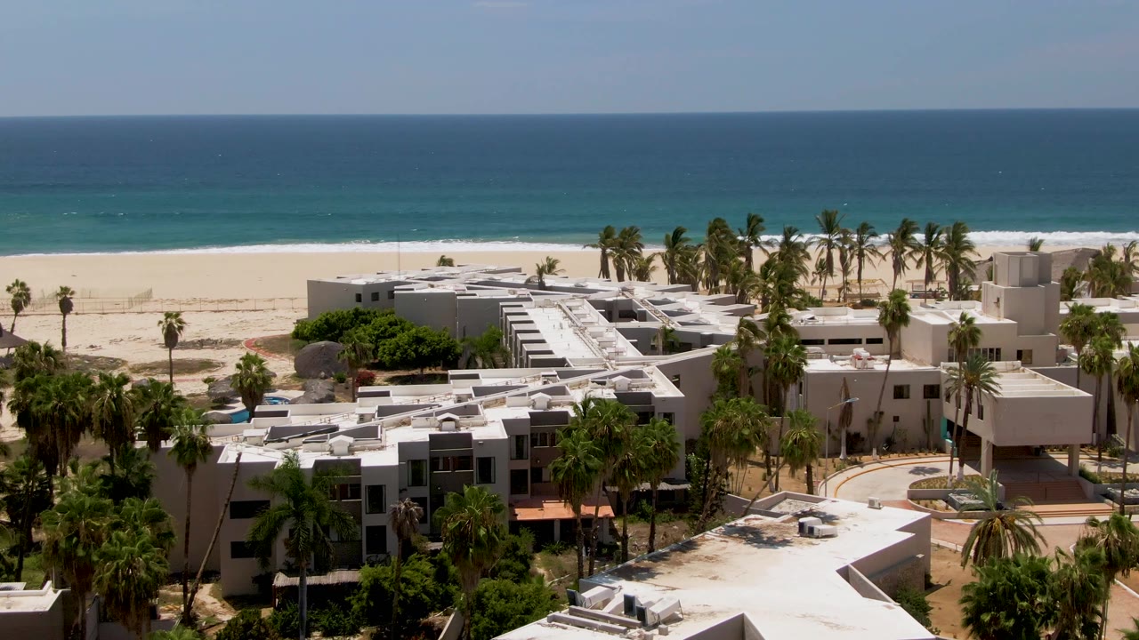 Resort complex, palm trees, sandy beach, and calm ocean under bright sun.