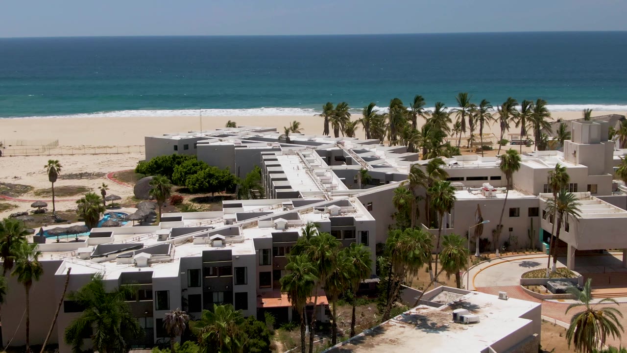 Aerial view in San Jose Del Cabo showing white resort rooftops, palm trees, a sandy beach, and blue ocean with no visible people.