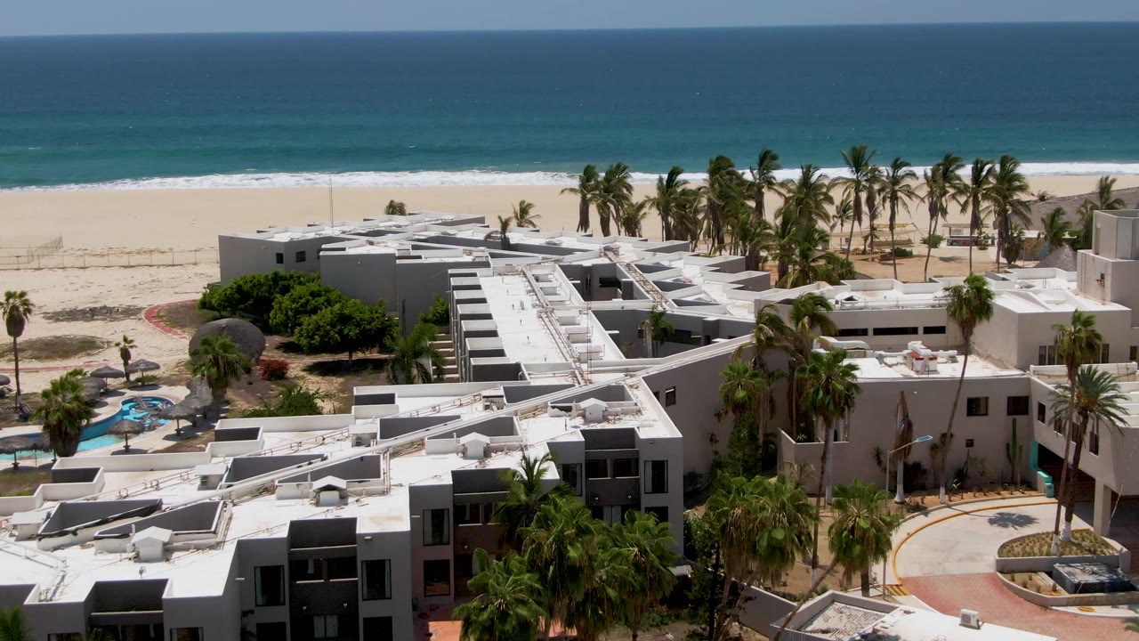 San Jose Del Cabo aerial view showing more of the beachfront resort complex, palm clusters, pale sand, and a dark-to-blue ocean horizon with no visible people.