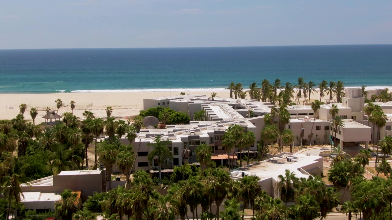 Aerial frame in San Jose del Cabo showing white resort rooftops and palm trees below a wide band of white sand and blue ocean, with no visible people.