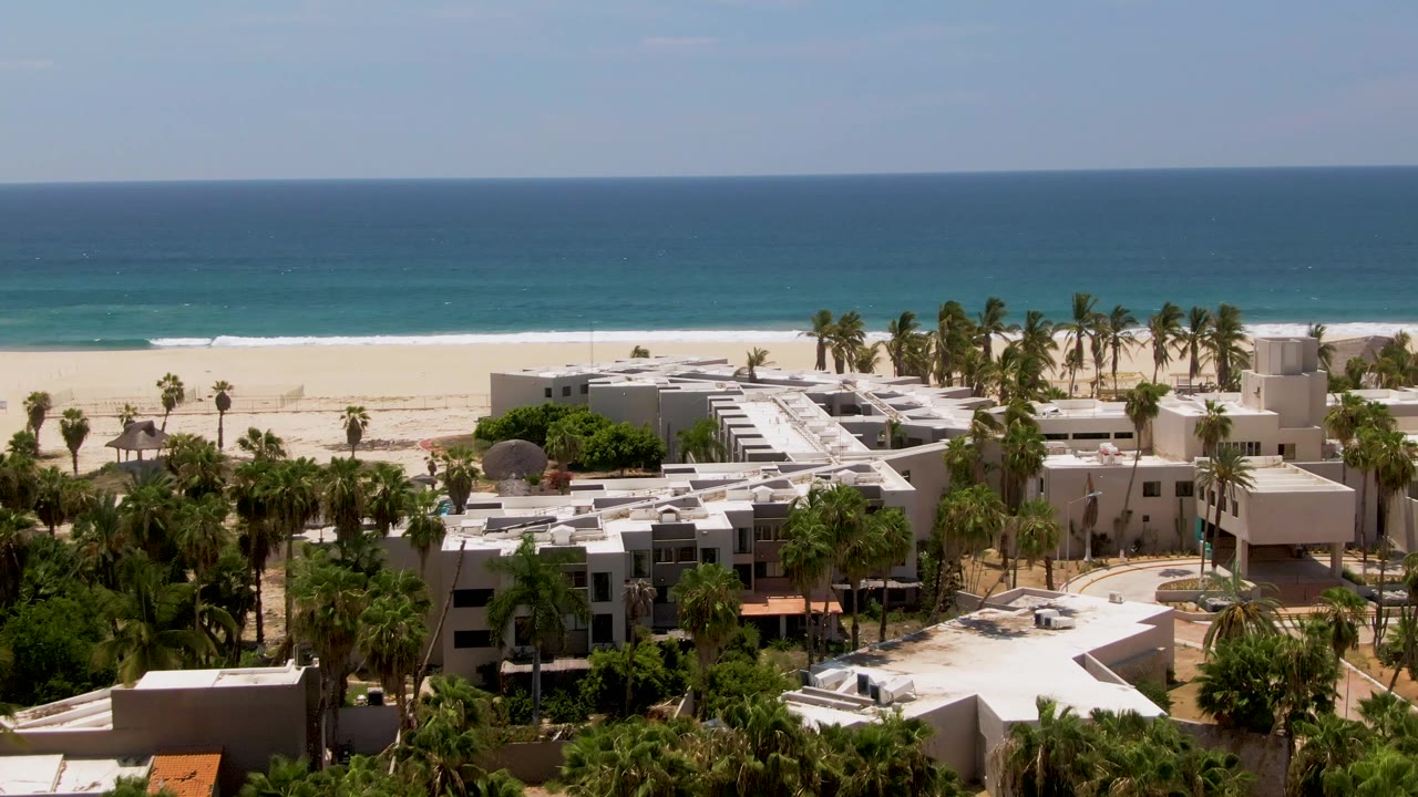 High aerial view over San Jose del Cabo resort buildings and palms, with the sandy beach and turquoise-to-deep-blue ocean stretching across the background, no visible people.