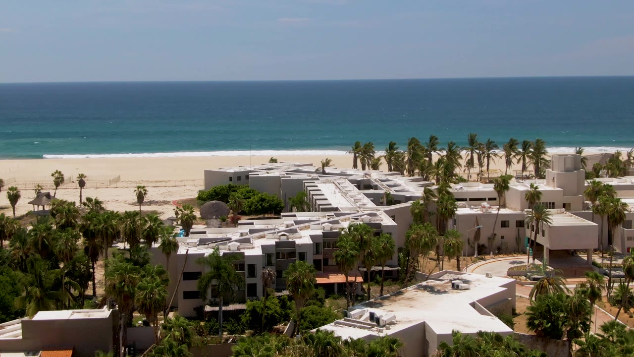 Drone-height view of a San Jose del Cabo beachfront resort, with white buildings and palms filling the lower frame and a bright sand beach and ocean behind, no visible people.