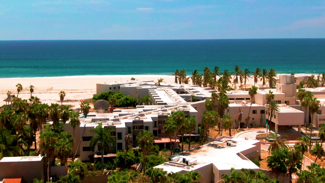 Elevated view in San Jose del Cabo showing white-roofed beachfront buildings, palm trees, pale sand, blue ocean, and no visible people at the start of the clip.