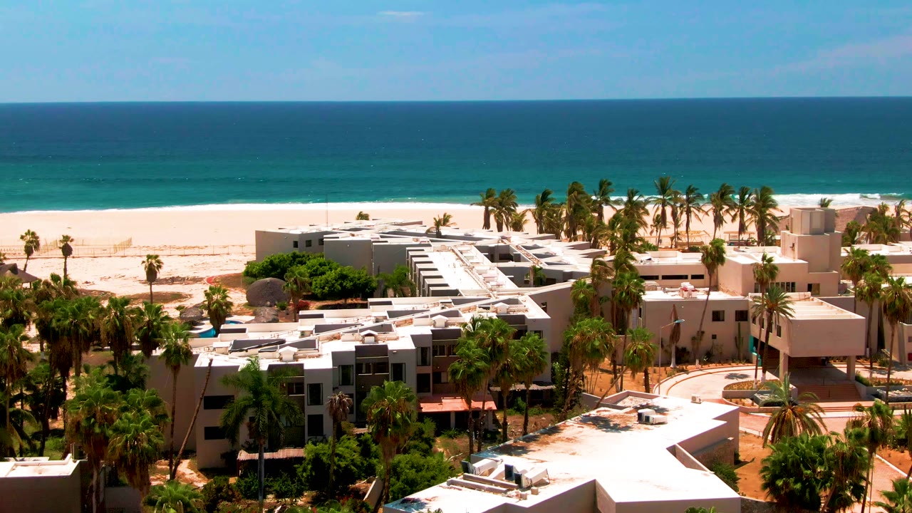 Elevated San Jose del Cabo frame with modern low-rise buildings, dense palms, a bright beach, calm blue ocean, and no visible people near the mid-run point.
