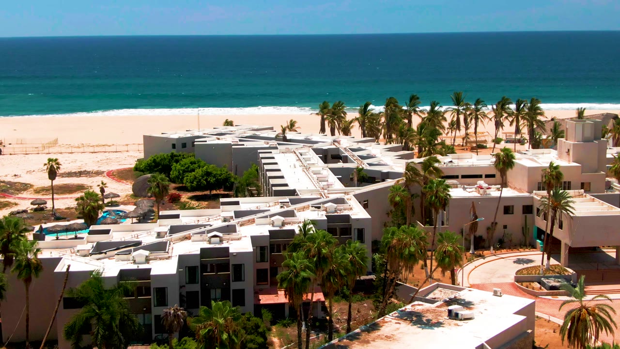 Aerial view of white beachfront resort buildings beside palms, sand, and blue ocean.