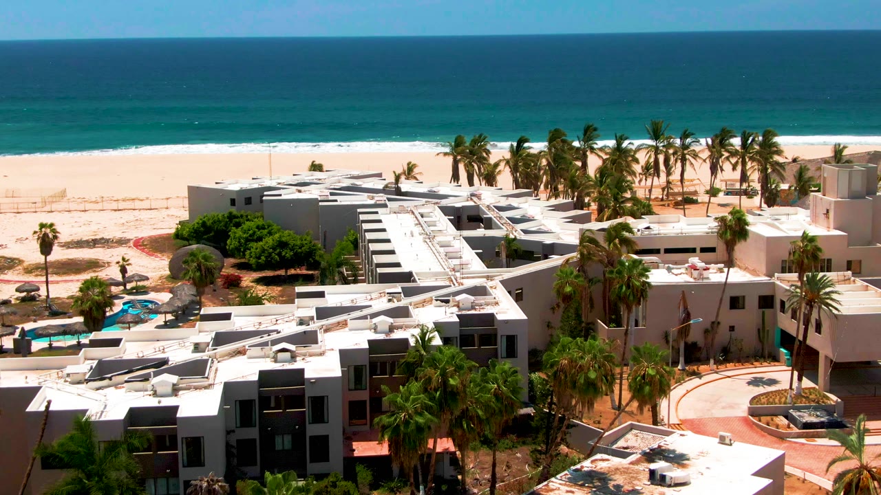 Elevated view of the San Jose del Cabo resort rooftops and palm-lined grounds, with the sandy beach and ocean horizon visible behind them, and no visible people.