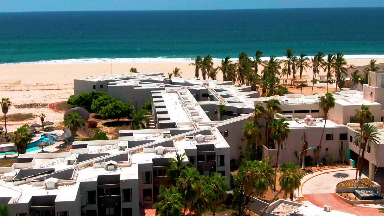 Aerial view of a San Jose del Cabo beachfront resort with white buildings, clustered palms, small blue pools, open sand, and no visible people.