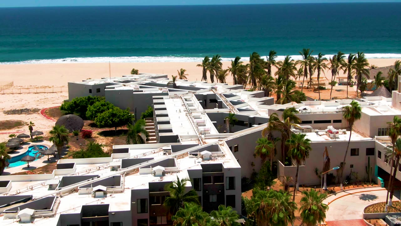 Closer elevated frame over white resort roofs in San Jose del Cabo, with palms crossing the midground, pale sand and blue water behind, and no visible people.