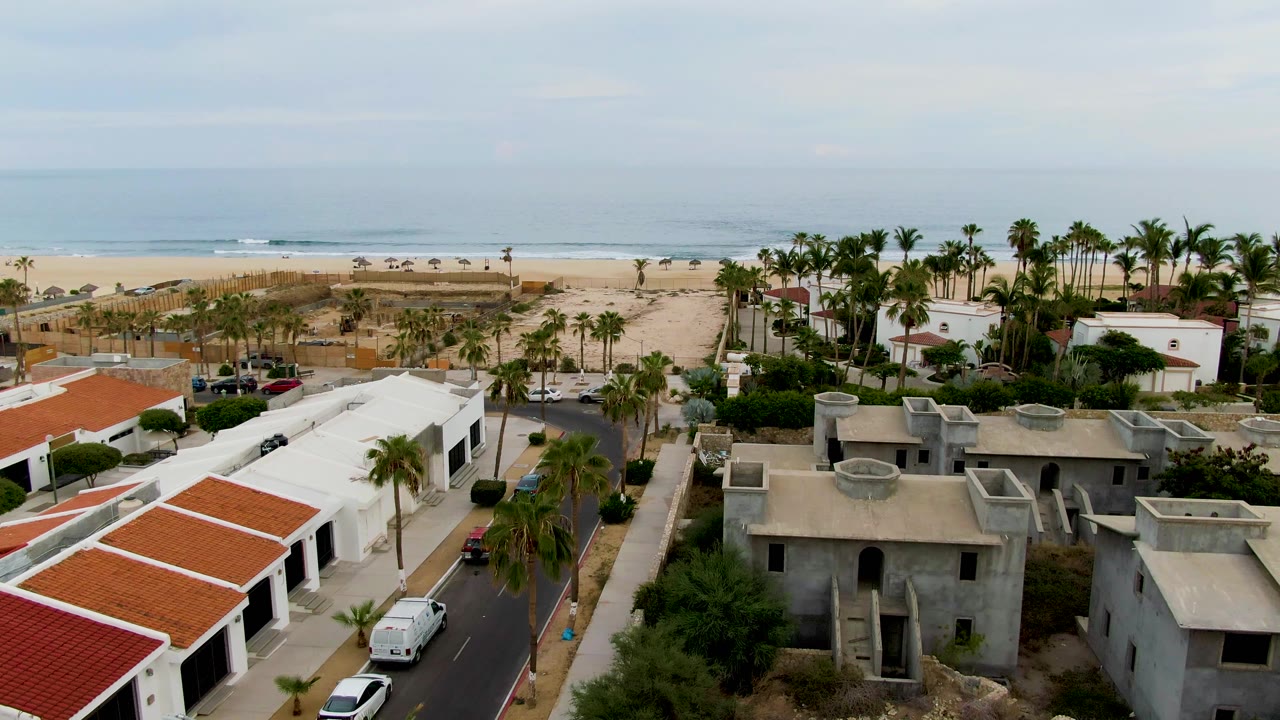 Aerial frame over San Jose del Cabo beachfront homes with red tile roofs, gray and white buildings, palm trees, a road, sand, ocean, and no visible people.