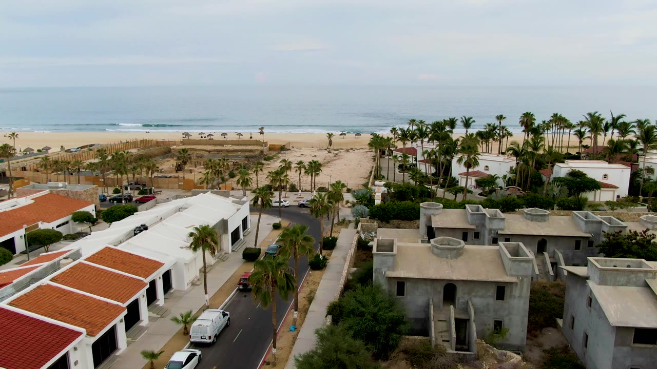 Drone frame in San Jose del Cabo showing more of the beachfront road, white residential buildings, palms, beach umbrellas, surf line, and no visible people.