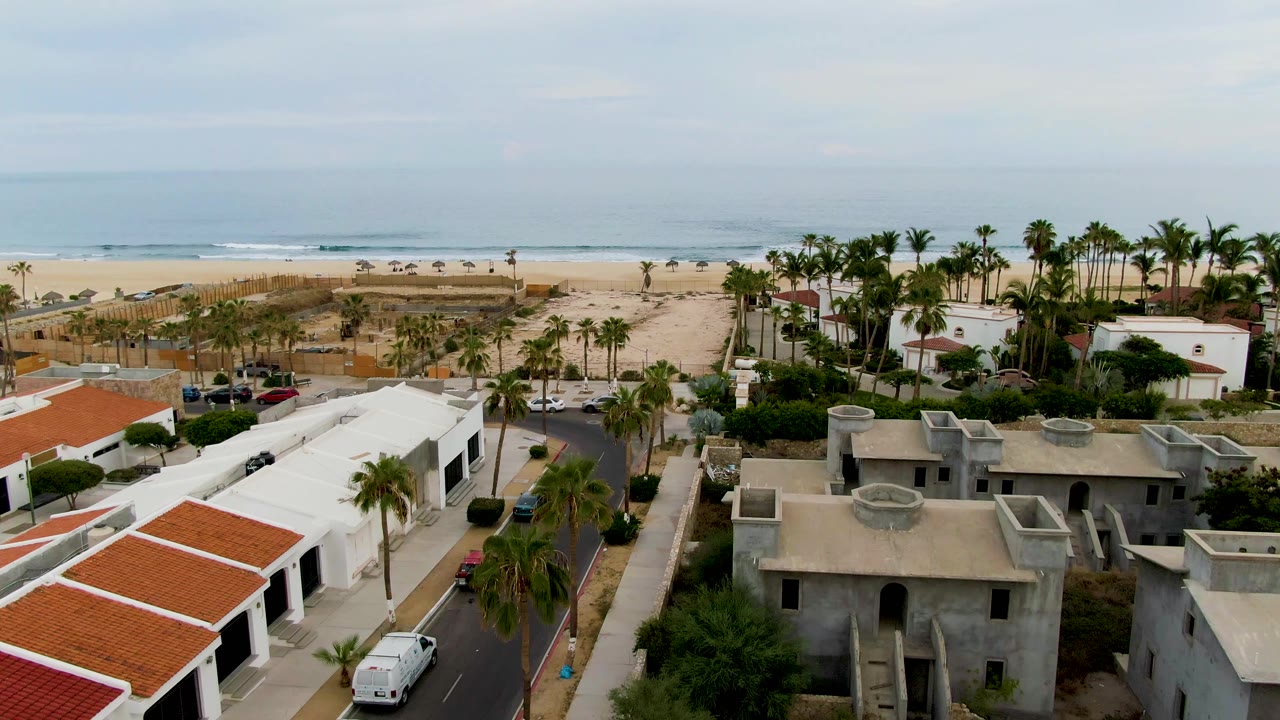 Aerial frame over San Jose del Cabo with a gray unfinished-looking building in the foreground, palms and parked cars along the road, sand, ocean surf, and no visible people.