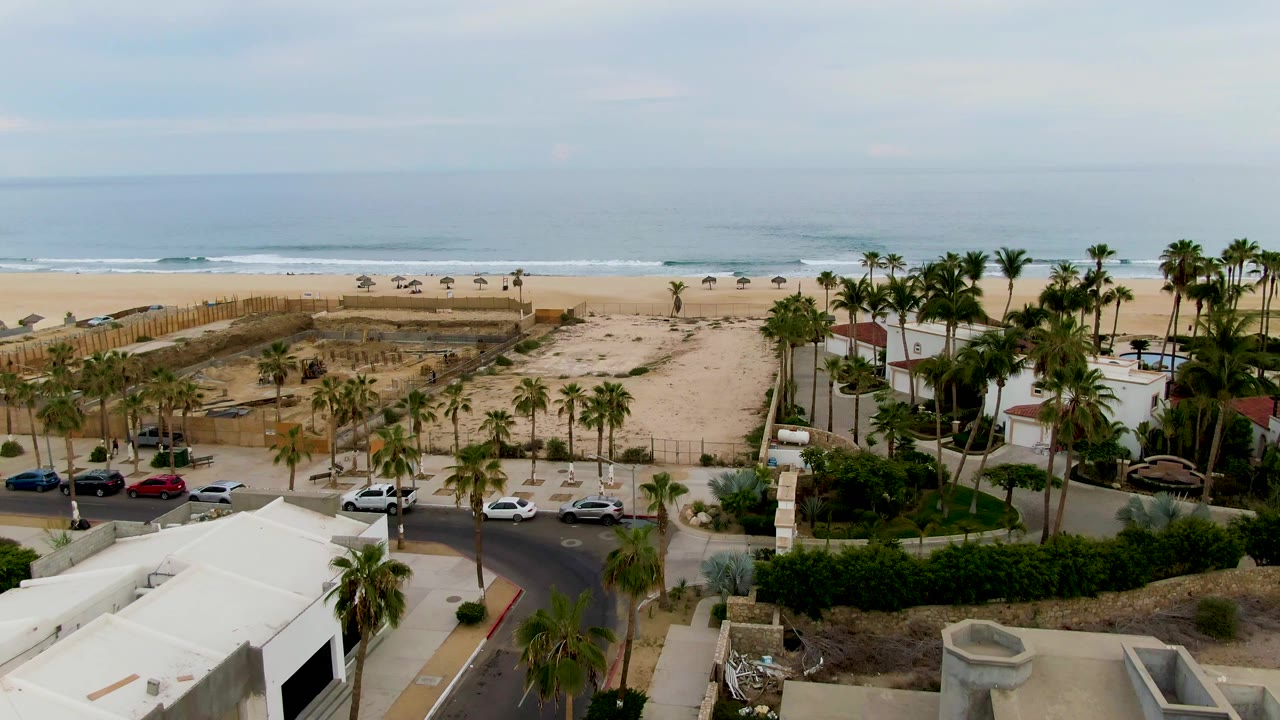 Aerial frame from San Jose del Cabo, Baja California Sur showing a sandy beach, palm-lined road, coastal buildings, parked cars, and no visible people.