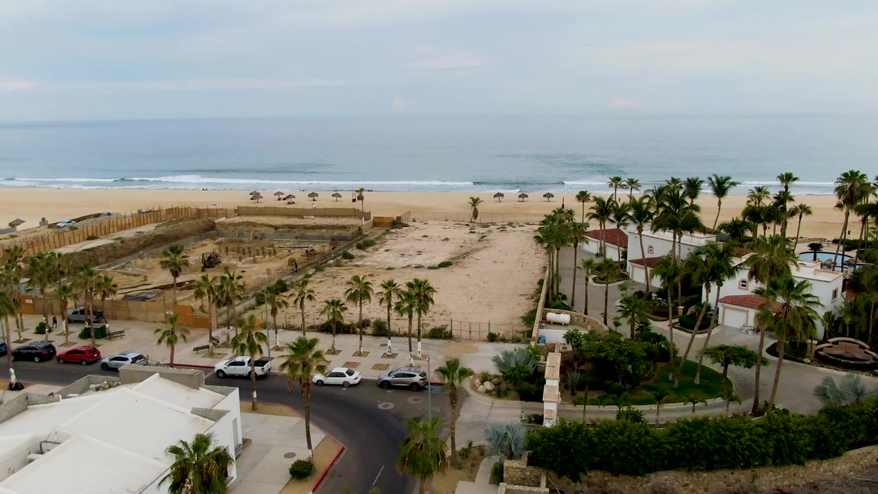 Elevated coastal view with palms, fenced sand areas, parked cars, buildings, and ocean surf.