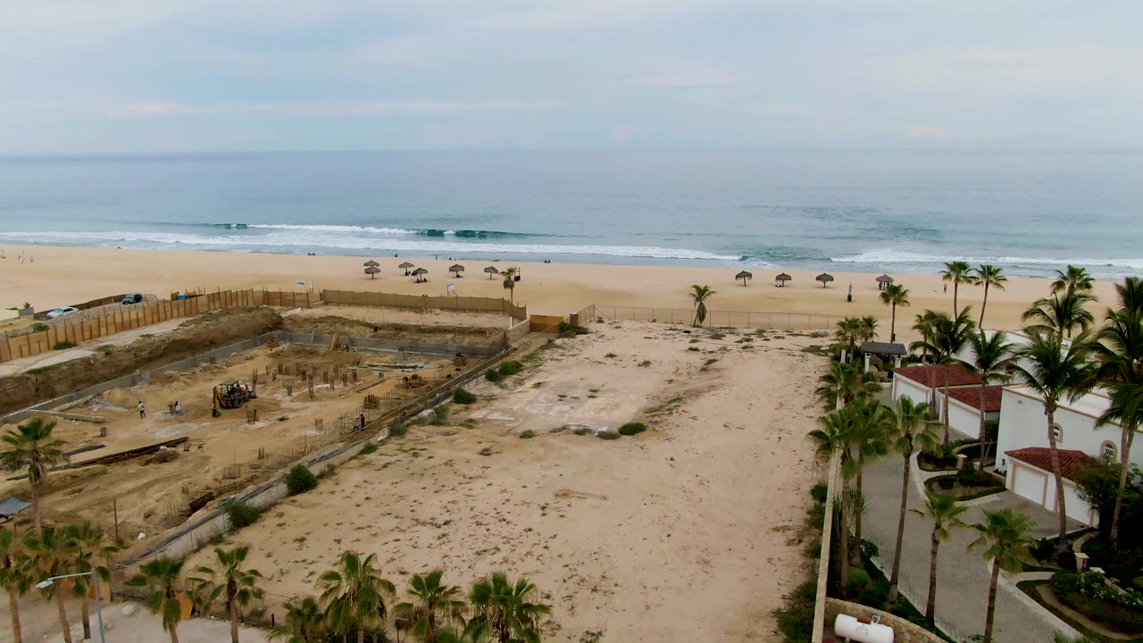Opening aerial frame over a sandy construction site in San Jose del Cabo, with palm trees, beachfront buildings, beach umbrellas, ocean waves, and no clearly visible people.
