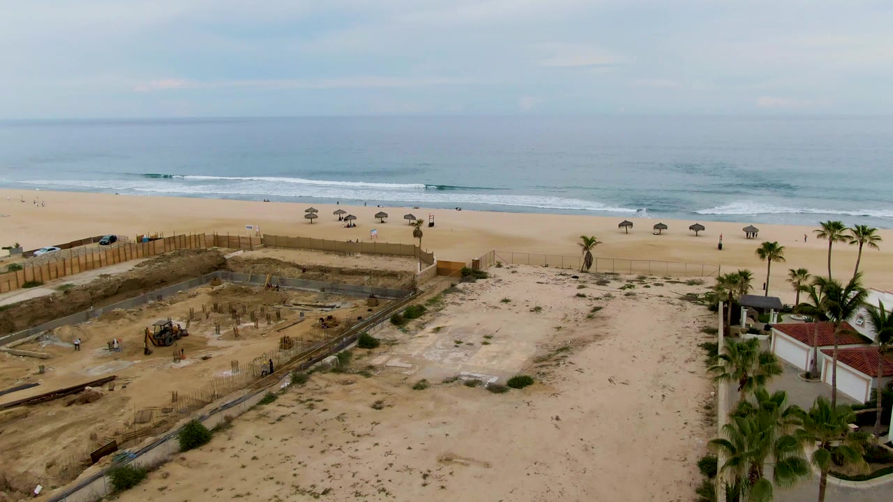 Aerial view over the San Jose del Cabo beach site, with a rectangular excavation area, scattered equipment, palms and red-roofed buildings at right, and surf breaking in the background.