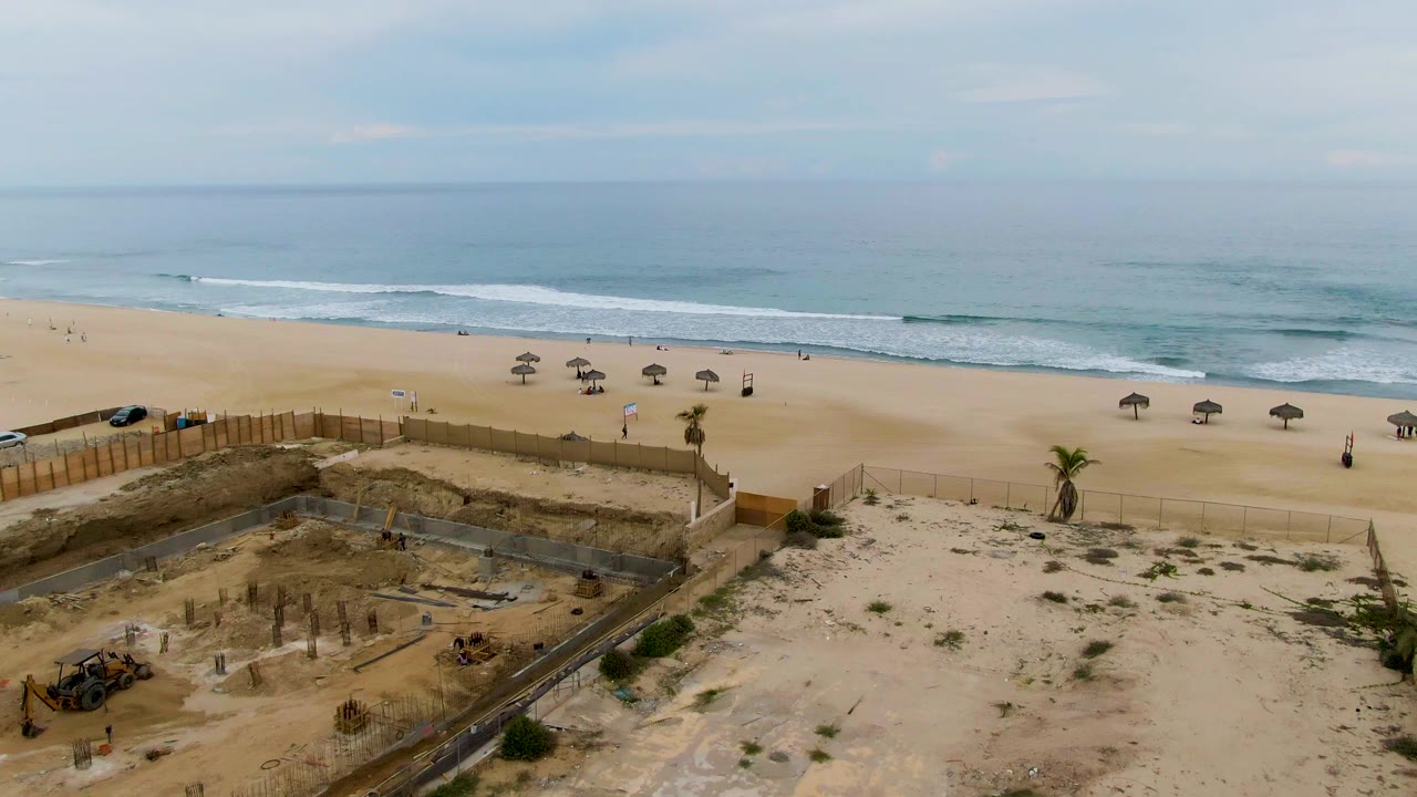 Aerial view in San Jose del Cabo showing a sandy construction area and fencing in the foreground, umbrellas and tiny beachgoers on the beach, and ocean waves beyond.