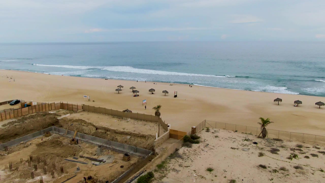 Wide aerial frame over San Jose del Cabo with a partially built structure in the foreground, tan beach sand, several umbrellas, distant beachgoers, and soft blue-gray ocean water.