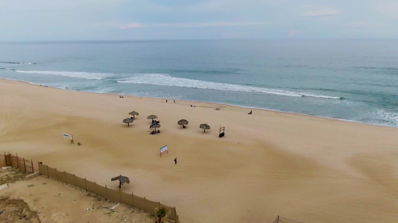 High aerial view over a sandy San Jose Del Cabo beach with a fence at lower left, several thatched umbrellas, small visible people, beach signs, and soft ocean waves beyond.